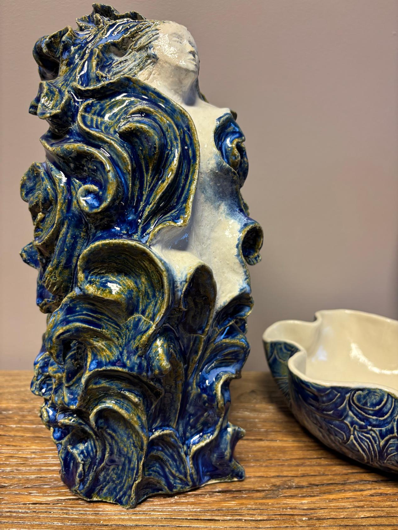 Håndlaget keramikkfigur med kvinneskikkelse omgitt av blå, bølgete former.